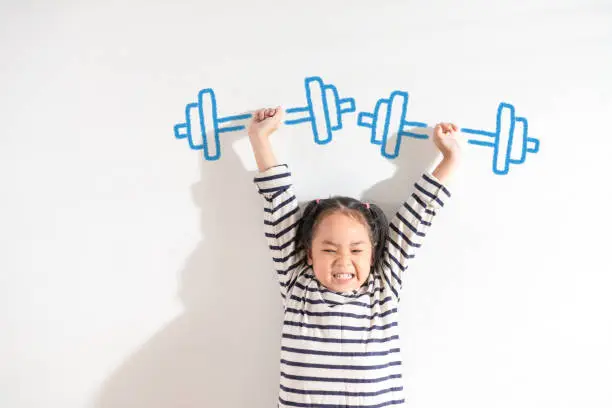 Photo of Funny Positive strong Asian little toddler kid girl lifting weight against the textured white background. For empowering women, girl power and feminism, sport, education, and creative future Ideas.