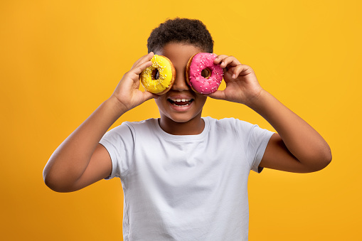Funny afro american preteen boy in white t-shirt holding colorful delicious donuts over eyes, cheerful black guy having fun while enjoying tasty sweets over yellow studio background