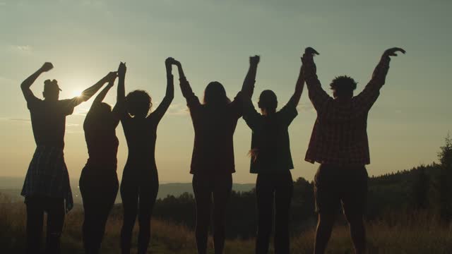 Silhouette of diverse multiracial friends standing with arms raised on mountain peak at sunset