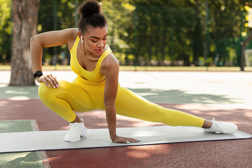 Sporty Healthy Lifestyle Concept. Confident determined African American lady in yellow top, leggings and white sneakers stretching leg muscles on yoga mat, doing side lunges in skandasana pose