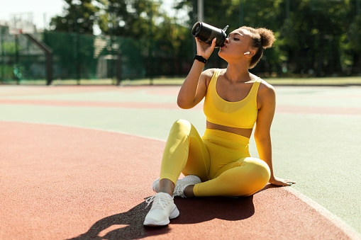 Energy, Vitality, Wellbeing Concept. Fit African American woman in wireless earbuds sitting on basketball field ground, holding shaker, drinking water or protein, taking break after outdoors exercise