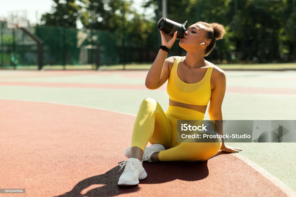 Portrait of sporty black woman in yellow sportswear drinking water Energy, Vitality, Wellbeing Concept. Fit African American woman in wireless earbuds sitting on basketball field ground, holding shaker, drinking water or protein, taking break after outdoors exercise Exercising Stock Photo