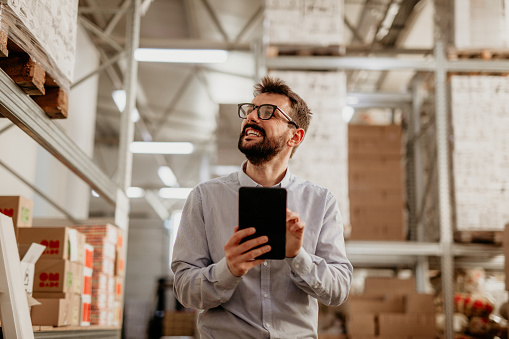 A spontaneous photo of a handsome bearded young adult man, with eyeglasses on, walking through the storage, while doing an online checkout on the tablet that he’s holding, facing up and typing with a smile on his face