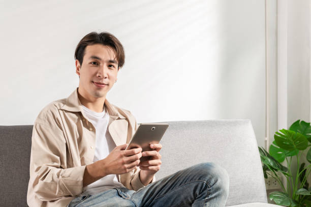 istock Happy smiling Asian student sits and relaxes on a couch using modern tablet browsing unlimited wireless internet, positive young man freelancer working uses wifi at a home apartment, technology idea. 1342241061