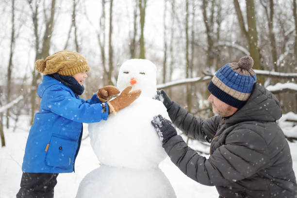 niño pequeño con su padre construyendo muñeco de nieve en un parque nevado. ocio activo al aire libre con niños en invierno. niño durante un paseo en un parque de invierno nevado - pareja acogido al aire libre fotografías e imágenes de stock