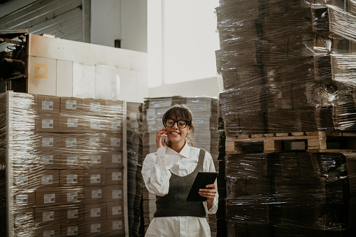 Spontaneous photo of a loving young adult woman, walking inside the warehouse, doing the shipment organization online, over the tablet while talking on the phone. She’s elegant casually dressed, looking like working with ease, smiling