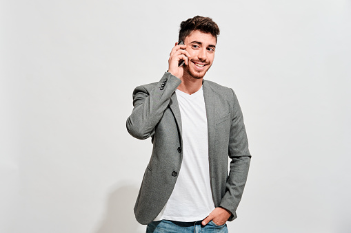 Happy young man wearing jeans, a white tshirt and a grey blazer talking with someone on his mobile phone