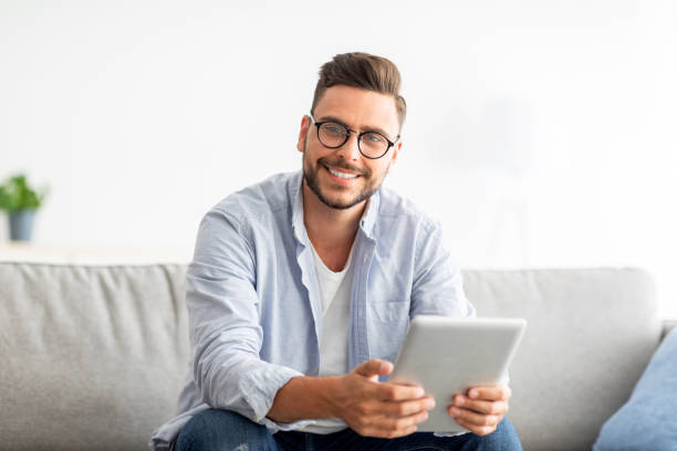 istock Technology concept. Excited millennial man using digital tablet, sitting on sofa at his modern apartment 1342242989