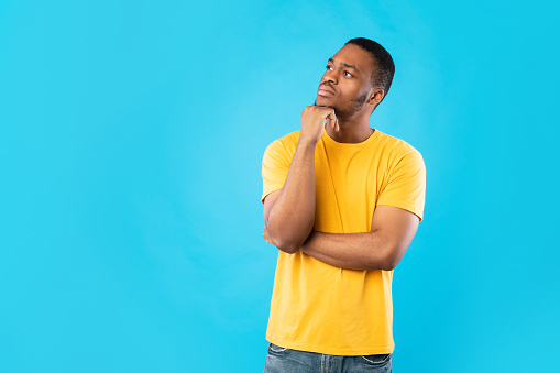 Pensive African American Man Thinking Touching Chin Looking Aside Posing Standing On Blue Background. Studio Shot Of Thoughtful Black Millennial Guy. Let Me Think Concept