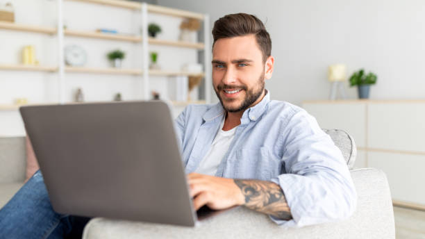 istock Positive guy reclining on sofa with laptop at home interior, urfing internet or scrolling social networks, panorama 1342243662