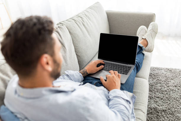 istock Advertisement concept. Young guy using modern laptop with blank screen, sitting on sofa, mockup, over shoulder shot 1342243990