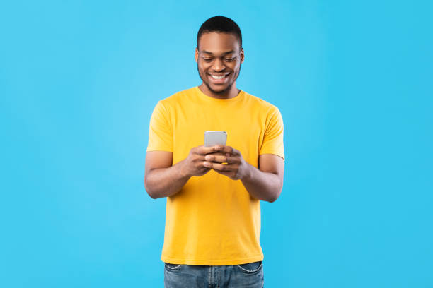 hombre afroamericano usando teléfono móvil sobre fondo azul de estudio - black sign holding vertical fotografías e imágenes de stock