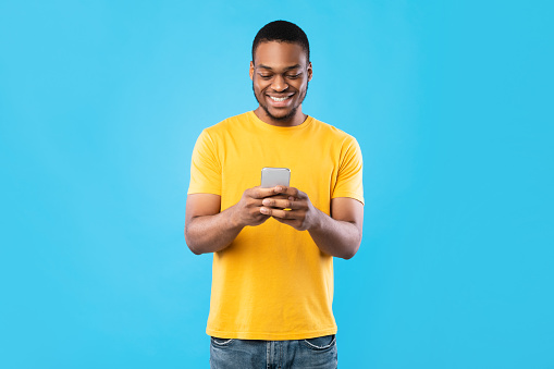 Happy African American Male Using Mobile Phone Texting Standing Over Blue Studio Background. Cheerful Black Guy Browsing Internet. New Application For Your Smartphone. People And Gadgets