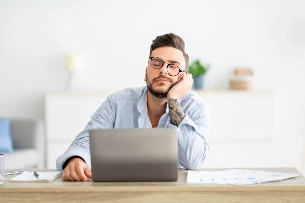 Overworked young freelancer man sitting at workplace and sleeping, napping while working on laptop computer, placed head on hand, feeling tired, copy space
