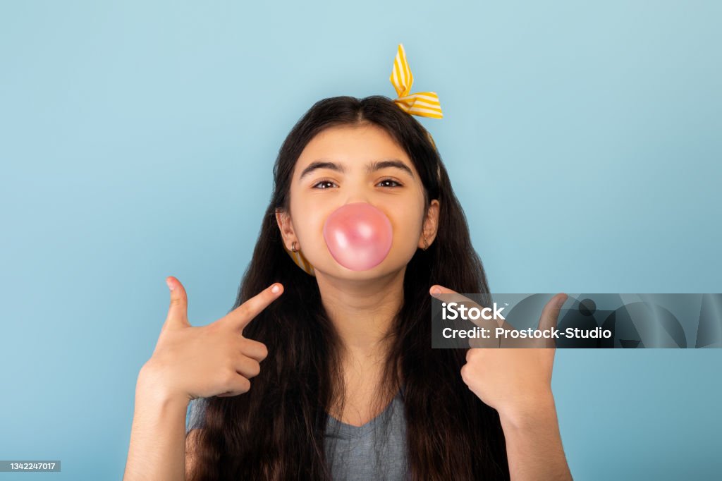 Funny Indian Teen Girl Blowing Bubble From Sugar Free Chewing Gum Pointing  At It Over Blue Studio Background Stock Photo - Download Image Now - iStock
