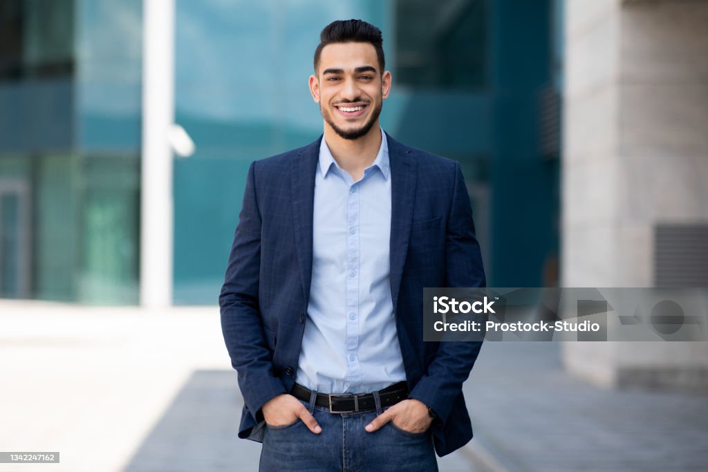Handsome middle-eastern guy businessman posing next to office center Handsome middle-eastern guy young businessman in stylish suit posing next to office center, smiling arab entrepreneur having break after successful busienss meeting, standing on street, copy space Businessman Stock Photo