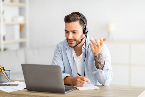 istock Web conference. Young freelancer man in headset making video call on laptop, sitting at workplace at home 1342247207