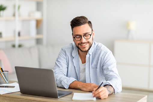 Happy millennial man looking for job online, sitting at table, using laptop and taking notes, living room interior, free space. Excited guy working online from home, remote job