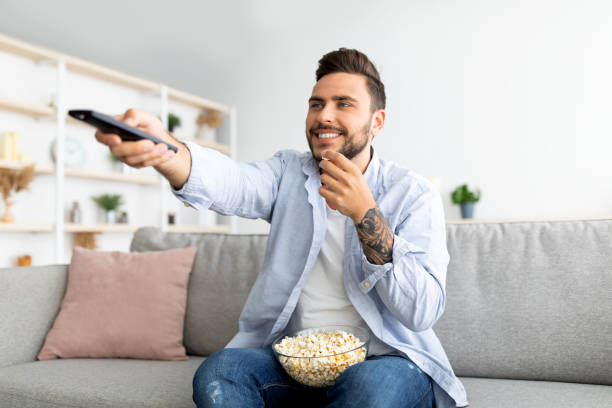 istock Cheerful millennial guy watching TV and eating popcorn, switching channels with remote controller 1342248096