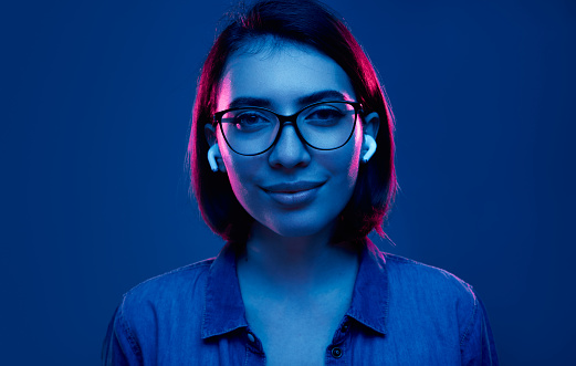 Positive young female in stylish eyeglasses listening to music through true wireless earbuds and looking at camera in studio with neon light