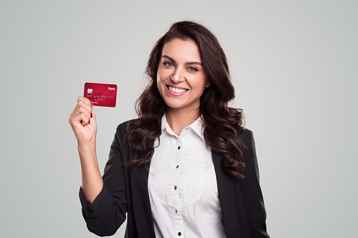 Positive adult businesswoman in formal clothes smiling friendly and demonstrating red credit card while recommending banking service against gray background