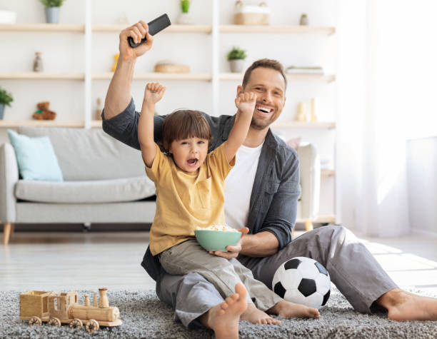 emotionaler kleiner junge, der die lieblingsfußballmannschaft anfeuert, zusammen mit papa sport schaut, zu hause auf dem boden sitzt - soccer child indoors little boys stock-fotos und bilder