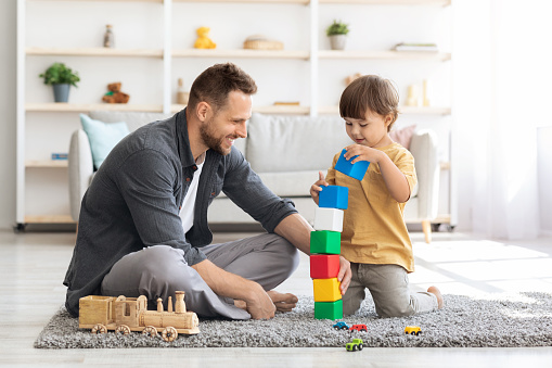 Playtime. Cheerful loving father playing colorful blocks with his little son, boy building big tower, spending free time together at home interior