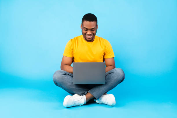 hombre negro feliz usando computadora portátil trabajando en línea sobre fondo azul - sentado fotografías e imágenes de stock