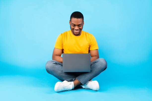 happy black man utilisant un ordinateur portable travaillant en ligne sur fond bleu - yellow and black photos et images de collection