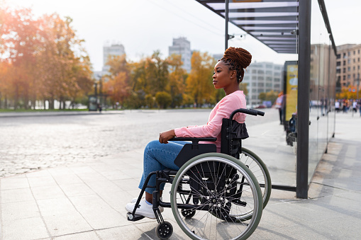 Lack of public transport for disabled people. Young black paraplegic woman in wheelchair feeling upset, waiting on bus stop in autumn, cannot board vehicle suitable for handicapped persons, copy space