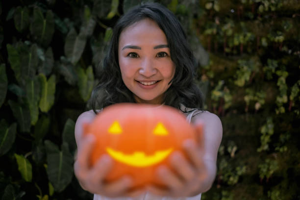 Close-up shot of beautiful smiling Asian woman standing in front of a living wall vertical garden while holding a Jack O'Lantern carved pumpkin.
