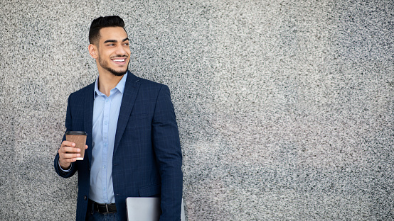 Handsome arab guy businessman in formal outfit with coffee to go and laptop standing outdoors next to grey building wall, looking at copy space, smiling, having break after successful business meeting