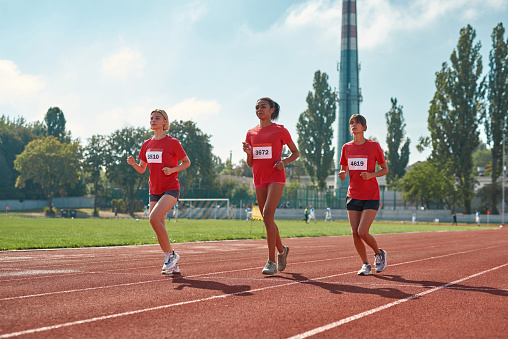 Full length shot of athletic young women running together during championship on track and field at the stadium on a summer day. Sports, race, motivation concept