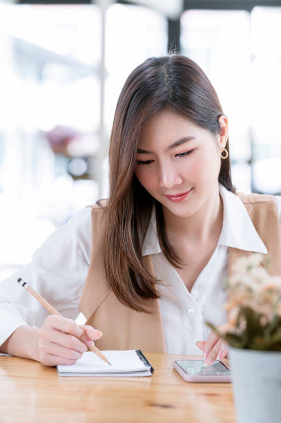 istock Portrait of young beautiful woman writing on notebook and using smartphone while sitting at office desk. 1342265303