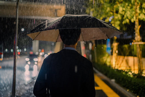 Asian businessman stands with an umbrella and using phone while waiting for a bus in the dark at the edge of the road.