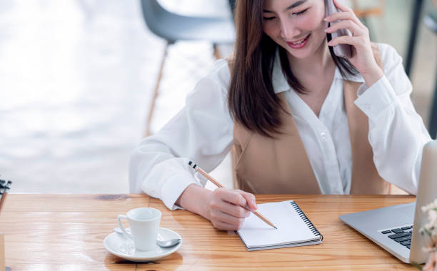 istock Cropped shot of woman hand writing on notebook while using smartphone and working on business at office. 1342270791