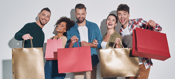 Group of young beautiful people in casual clothing carrying shopping bags and smiling while standing against gray background