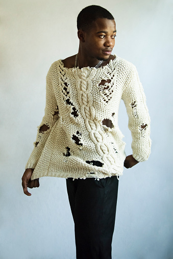 Young adult man with dark skin studio portrait. He is standing, with black pants and a custom made wool sweater, silver rings and necklace. Vertical indoors waist up studio shot in natural light with copy space.
