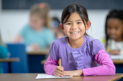 A group of multi-ethnic elementary students each sit at their desks in class with pencils inn hands as they work away on a writing assignment.  The focus is on a sweet girl of Asian decent, dressed in a sweater with stars on it, who is looking up from her page to smile.