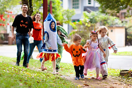 Child in Halloween costume. Mixed race Asian and Caucasian kids and parents trick or treat on street. Little boy and girl with pumpkin lantern and candy bucket. Baby in witch hat. Autumn holiday fun.