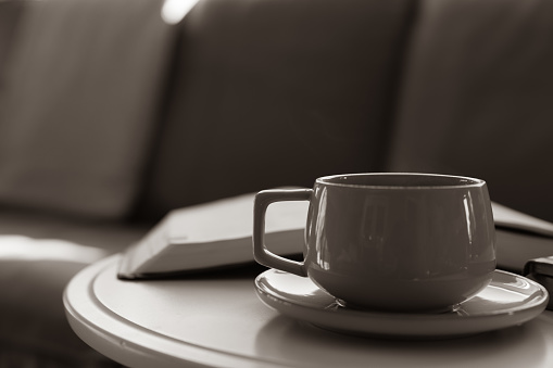 Close up of a cup of coffee on a saucer and open book on a small round white table sitting in front of a couch