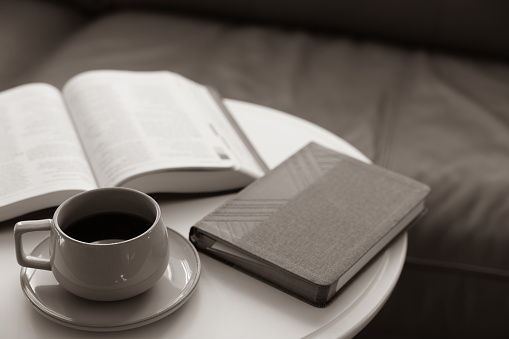 Cup of coffee on saucer with a closed devotional book and an open bible sitting on a round white side table in front of a sofa with copy space