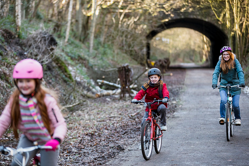 A front-view shot of a mother with her young boy and girl riding their bikes along a footpath, they are wrapped up in warm clothing on a winter's day. The young girl is leading the way.