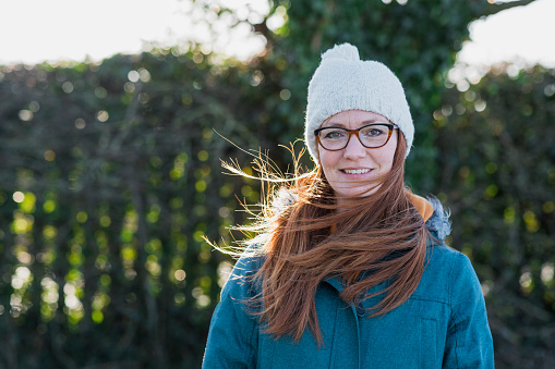 A front view headshot portrait of a mid-adult caucasian woman standing outdoors getting some fresh air on a winter morning, she is wrapped up in warm clothing on a winter's day. She is smiling and looking at the camera.