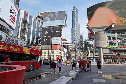 Toronto, Canada - August 30, 2021: Large commercial signs tower over the city traffic and pedestrians  at Yonge-Dundas Square. Travellers wait outside a double-decker tour bus on a summer morning.in the Church-Yonge Corridor.