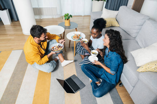 Three young carefree friends two women and one man sitting in the apartment on the floor listening to music on a laptop eating and having fun for a free weekend without worries Three young carefree friends two women and one man sitting in the apartment on the floor listening to music on a laptop eating and having fun for a free weekend without worries roommate stock pictures, royalty-free photos & images