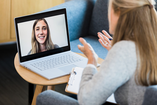 A female counselor gestures while talking with a young woman during a virtual therapy session.