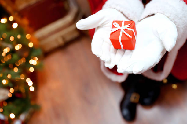 Red Christmas gift box in Santa hand. Giving present near decorated tree with lights on New Year's Eve.  Copy space