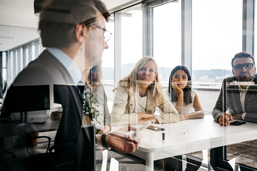 A group of smartly dressed co-workers are participating in a meeting. They are focused and concentrated. They are sitting behind a tall white desk in a bright modern office with large windows. Horizontal daylight indoor photo.
