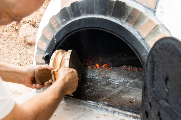 White painted artisan wood-fired oven built on the outside with the door open and man's hands blowing the embers with a bellows